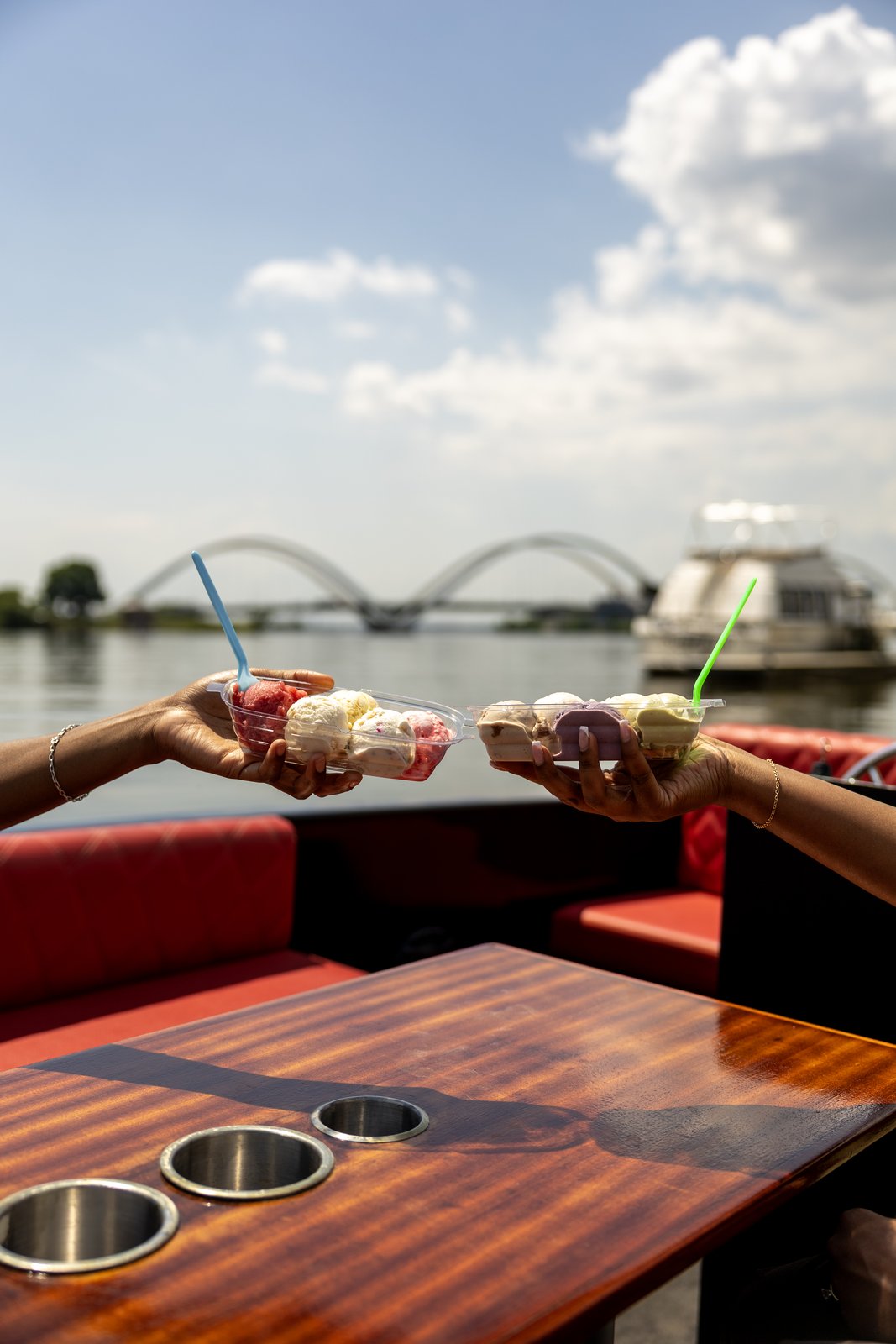 Two guests toasting Ice Cream Jubilee sample flights aboard a Yachtsy cruise, the Frederick Douglass Memorial Bridge on the Navy Yard waterfront behind