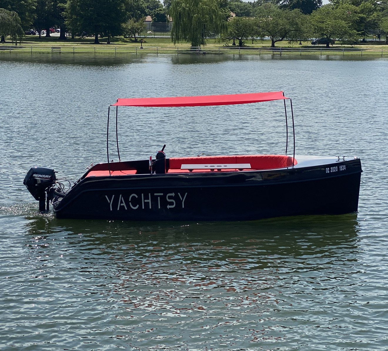 The Yachtsy picnic boat underway on the DC Tidal Basin — black hull, red bimini canopy, lush green trees